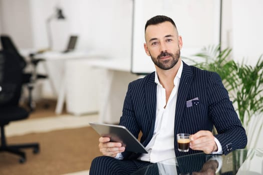 Confident positive european adult man in suit with beard uses tablet to manage business, drinks cup of coffee