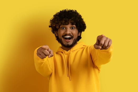 Amazed handsome indian guy pointing at camera, isolated on yellow