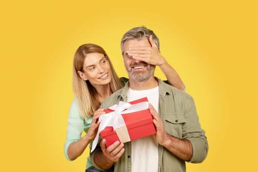 Happy european woman closing eyes to husband, giving gift box and celebrating birthday, posing on yellow background