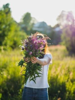 Young woman holds wild flowers bouquet outdoors in beautiful backlight sunset light, near green field in countryside. Summer, rural life and naturecore aesthetic concept
