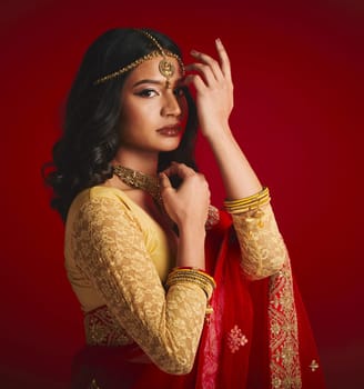 Portrait, celebration and Indian woman with fashion, traditional dress and jewellery against a red studio background. Face, female person or model with cultural clothes, religion and beauty with gold