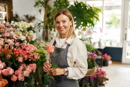 Smiling woman florist hold rose ready to sell on floral background and looking at camera
