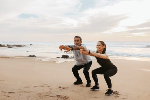 Happy senior caucasian couple in sportswear squats, enjoy training together on beach in morning, outdoor, full length. Health care, relationships, sports teamwork and active lifestyle