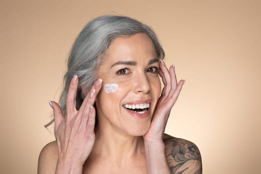 Portrait of beautiful senior grey-haired woman with moisturizer cream on her cheeks, testing new beauty product