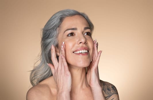 Beautiful senior woman touching her smooth face skin, applying moisturizer cream or lotion on cheeks, beige background