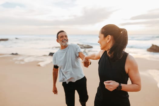 Laughing senior caucasian lady in sportswear leads man by hand, motivate for training and active lifestyle on beach in morning, outdoor. Jogging, relationships, sports together