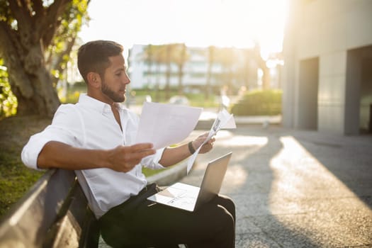 Man business analyst, accountant sitting on bench with laptop, holding papers, checking correspondence outdoors