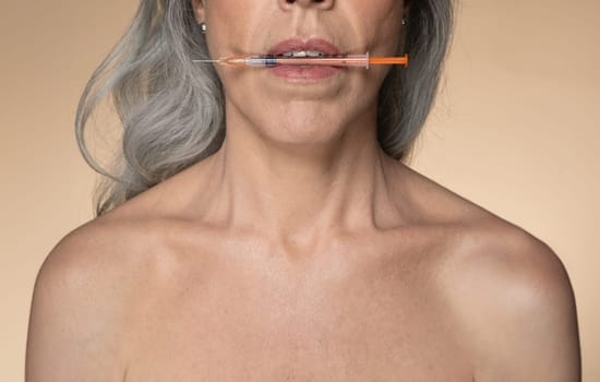 Cosmetology for aged women. European senior woman holding syringe with hyaluronic acid for lip augmentation in her mouth