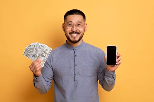 Cheerful Young Asian Guy Demonstrating Blank Smartphone And Dollar Cash Money Banknotes