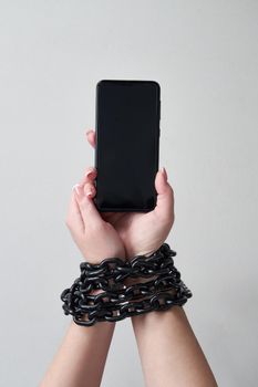 Iron chain that ties together hand and smartphone in concept of social media and internet addiction on gray background