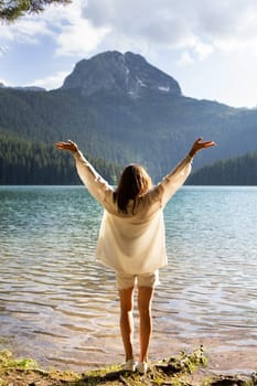 The girl stands with her back near a mountain lake in the background. Positive young woman traveling on a blue lake outdoors, adventure travel.