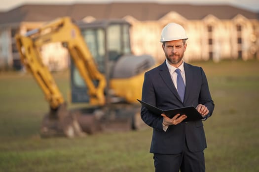Architect at a construction site. Architect man in helmet and suit at modern home building construction. Architect with a safety vest and suit. Confident architect standing at house background.
