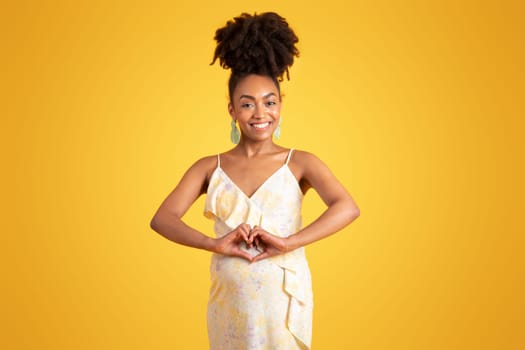 Glad millennial african american lady with belly making heart sign with hands, isolated on yellow background