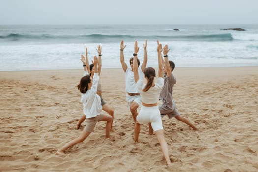 Group of young men and women in sportswear doing yoga on ocean shore, standing on mats in circle, practising asana exercise Virabhadrasana Warrior I Pose