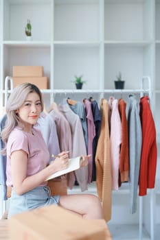 Portrait Of Asian Female Owner Of Fashion Store Checking Stock In Clothing Store With using notebook successful happy smile at small business, sme or ecommerce concepts