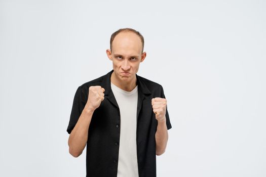 Defending himself stands in a stance with ready fists young bald man in black shirt with white t-shirt under. Angry young adult man showing he is ready for fight isolated on white background