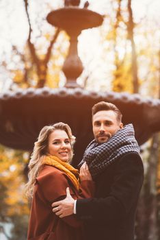 Half-length portrait loving couple of young people standing embracing and looking at camera with an old fountain on background at the autumn park wearing autumn coats. Toned photo