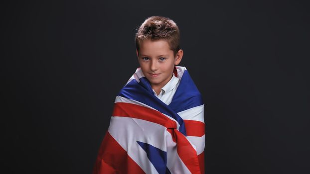 Little boy patriot standing side turned wrapped in a UK or British flag celebrates independence day expresses patriotism isolated on black background