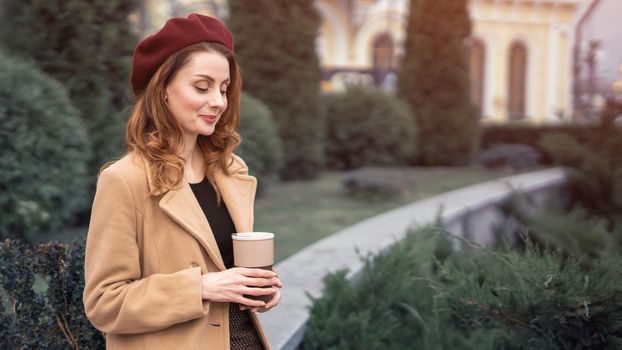Beautiful young woman holding a mug with coffee standing outdoors. Portrait of stylish young woman wearing autumn coat and red beret outdoors. Autumn accessories