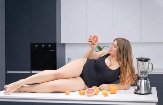 Sexy overweight girl laying on the kitchen table. An obese young sexy chubby white girl in black swimsuit at modern kitchen with fruits next to her