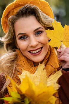 Close up. Young woman in beige knitted beret with autumn leaves in hand and fall yellow garden or park. Beautiful smiling young woman in autumn leaves