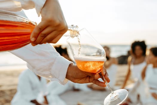 Lady pouring rose wine into glass while group of women having picnic on background on the beach, selective focus