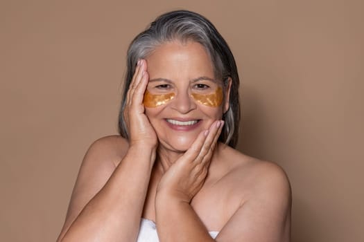 Cheerful elderly caucasian lady with gray hair enjoys anti-aging treatment, applies eye patches