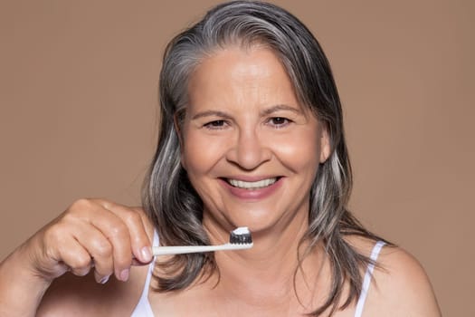 Cheerful elderly caucasian lady with gray hair brushes her teeth, isolated on brown background, studio