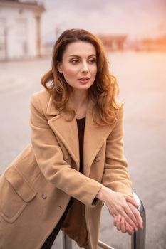 Beautiful Parisian woman standing outdoors posing leaned on handrail for camera in an autumn beige coat, beautiful hair with urban city background. Vertical portrait. Warm tinted photo