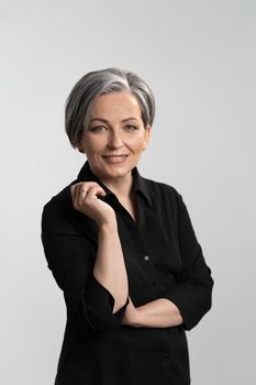 Grey haired mid aged pretty woman looking at camera with arms folded wearing black shirt isolated on grey background. Confident mature grey haired woman. Human emotions, facial expression concept