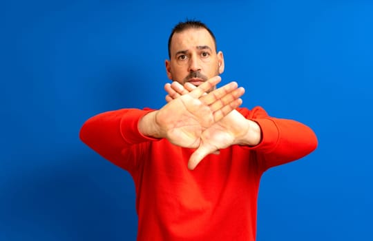 Bearded hispanic man in his 40s wearing a red jumper making a stop sign with his palms crossed in a reject and denial gesture, isolated on a blue background.