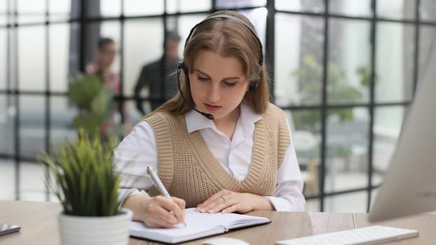 Attractive confident business woman, office manager, wearing headset using laptop while making, writing financial repor