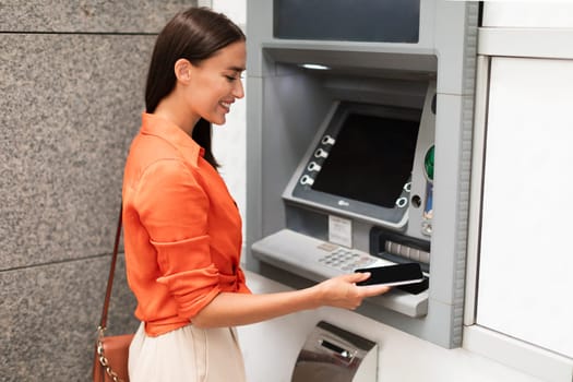 Lady using smartphone gaining access to ATM withdrawing money outside