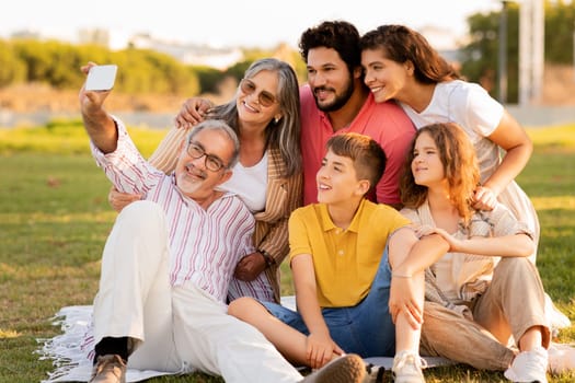 Positive caucasian multi-generation people rest, enjoy picnic in park, take selfie on smartphone