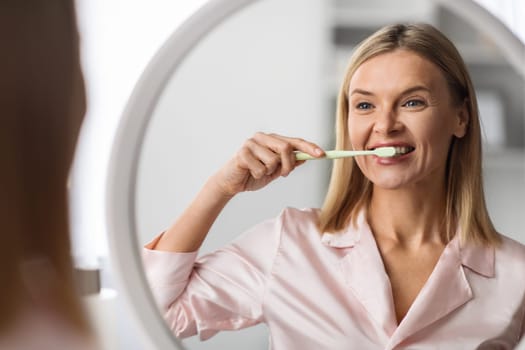 Beautiful Mature Female Brushing Her Teeth With Toothbrush Near Mirror At Home