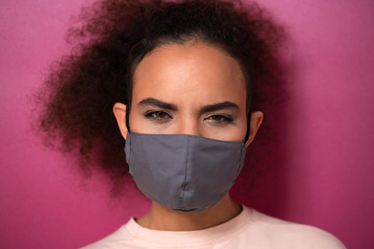 Questioning or studying look wearing reusable face mask African American charming girl in peachy t-shirt, to prevent others from corona COVID-19 and SARS cov 2 infection isolated on pink background