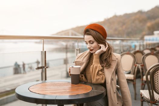 Thoughtful young woman looking at cup of coffee wearing red beret sitting on the terrace of a restaurant or cafe on background of an autumn urban city