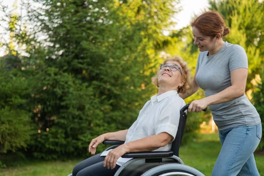 A caucasian woman is carrying an elderly mother sitting in a wheelchair through the park. Walk outdoors.