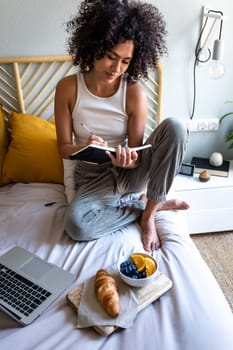 Multiracial latina woman writing daily affirmations on journal while eating breakfast in bed next to laptop.Vertical.