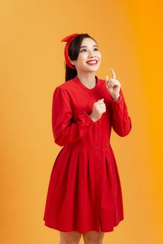 Surprised beautiful young woman in red dress is pointing. Three quarter length studio shot on orange background.