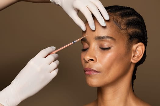 Beautician hands making hyaluronic acid injection, black middle aged woman getting cosmetic injection between eyebrows