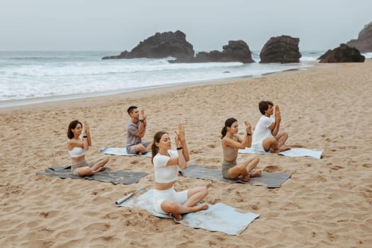 Calm relaxed group of women and man doing yoga on fitness mats, training in the morning on sandy beach, stting with crossed legs and doing namaste pose