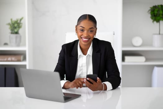 Portrait of black female entrepreneur sitting at workplace with laptop and with phone in hands, smiling at camera