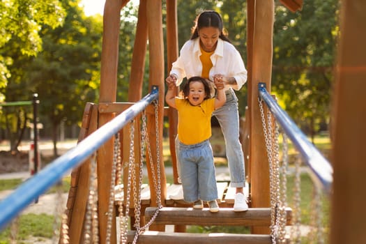 Asian Mother With Infant Daughter Walking Swinging Bridge On Playground