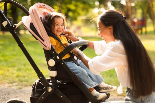 Happy Asian Mother Making Toddler Daughter Comfortable In Stroller Outdoor