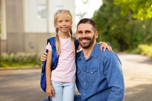 Back to school concept. Little girl with school bag embracing father near school, smiling together at camera