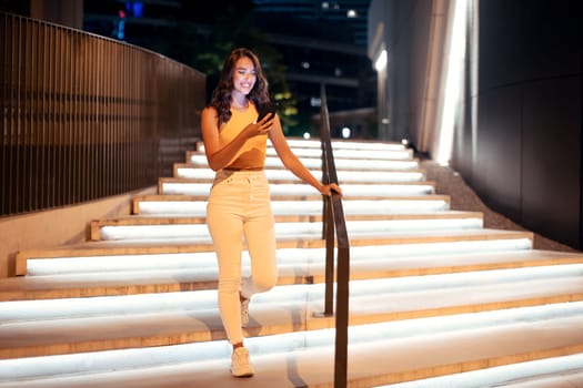 Full length shot of woman using phone during night walk in the city, going down the illuminated stairs, free space