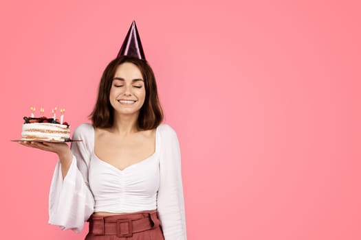 Satisfied millennial caucasian brunette woman in hat make wish with closed eyes, hold cake with candles