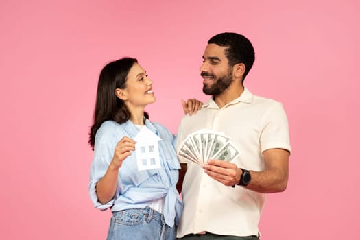 Cheerful millennial couple buying house, holding money cash, pink background
