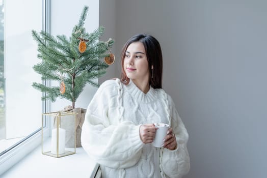 Merry Christmas and Happy New Year. Woman in warm white winter sweater standing next to the window at home at christmas eve holding cup with marshmallows, fir tree behind